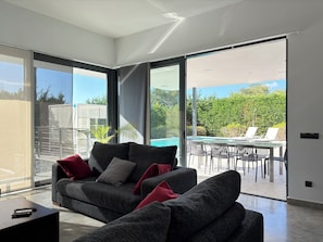 A modern living room with a grey sofa, a coffee table, and a view of an outdoor pool area. - Luxury Ibiza Villa – Walk to Beach in 3 Minutes, Pool & Outdoor Living (Cala Llenya)