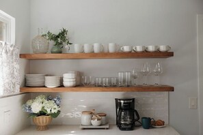 Una cocina con dos estantes de madera que sostienen tazas y platos blancos, una cafetera y una cesta con flores.