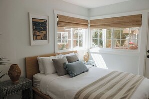 Un dormitorio con cama, almohadas, una mesita de noche, una obra de arte en la pared y una ventana con vistas al exterior.