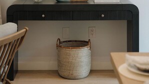 Ein Zimmer mit einer schwarzen Konsole, einer Vase mit grünen Blumen, einem gerahmten Bild, einem Korbwesen und einem Sessel.