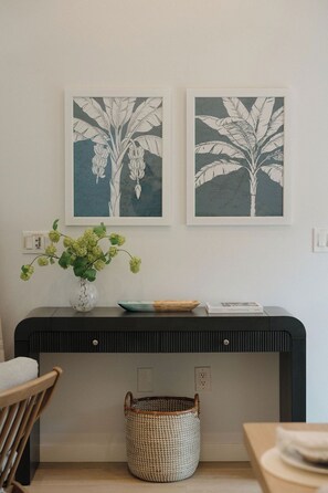 Ein Zimmer mit einer schwarzen Konsole, einer Vase mit grünen Blumen, einem gerahmten Bild, einem Korbwesen und einem Sessel.