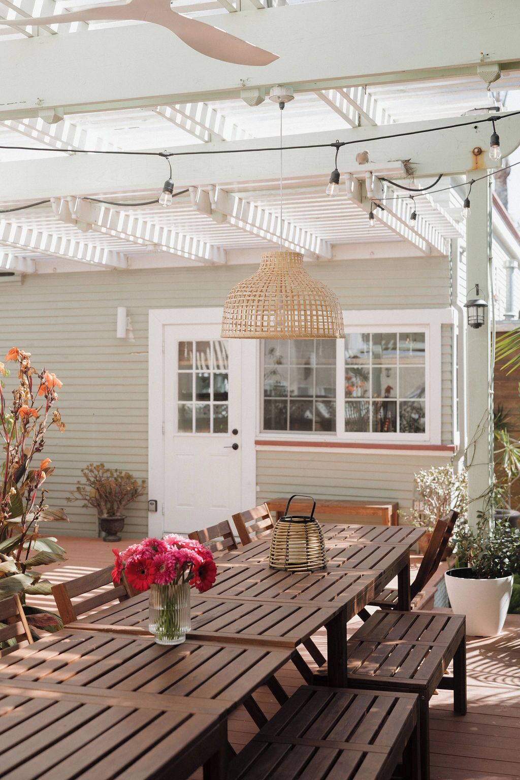 Une terrasse extérieure avec des tables et des chaises en bois, un luminaire suspendu en fibres tressées et des plantes en pot.