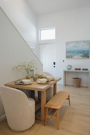 A dining area with a wooden table, chairs, and a bench. There is a window with a view of the outdoors, a door, and a painting on the wall. - Heart of OB, Walk to Beach, Townhome with Garage and Patio (San Diego)