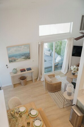 A modern living room with a large window, a painting, a sofa, and a dining area with a table set for two. - Heart of OB, Walk to Beach, Townhome with Garage and Patio (San Diego)