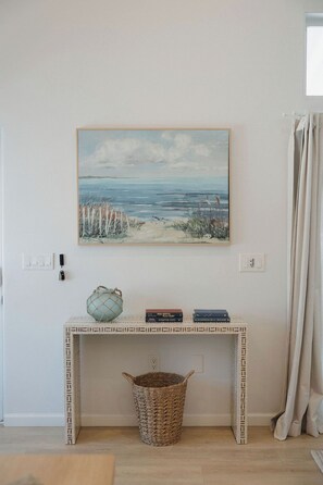 Una habitación con un cuadro de playa, una consola con libros y una cesta, y una ventana con cortinas.