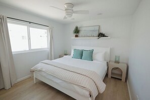 Un dormitorio con cama, ventilador de techo, ventana con cortinas, mesita de noche con lámpara y estante en la pared con una planta.