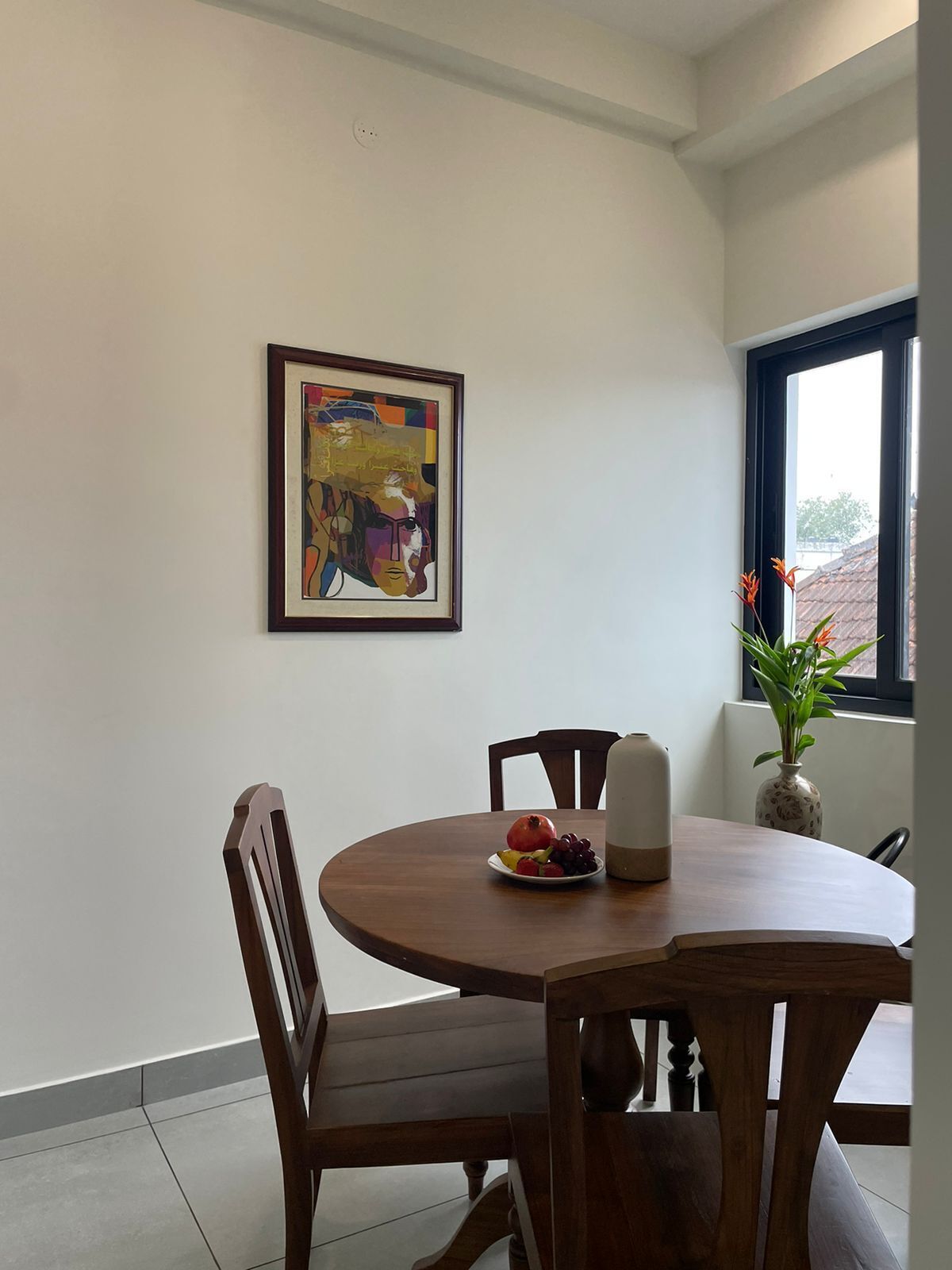 A dining area with a round table, chairs, a framed artwork, a vase with flowers, and a window with a view.