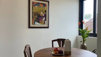 A dining area with a round table, chairs, a framed artwork, a vase with flowers, and a window with a view.