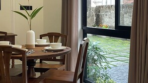 A dining area with a round table, wooden chairs, a vase with a plant, and a view of a garden through a large window.