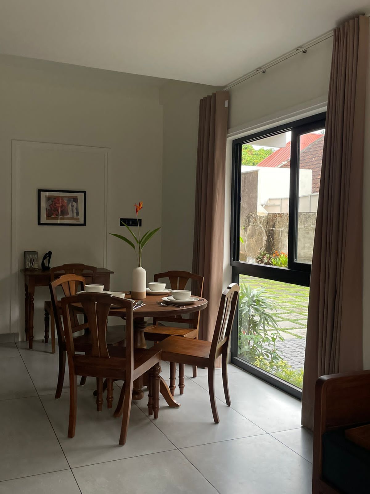 A dining area with a round table, chairs, a vase with flowers, and a window with curtains.