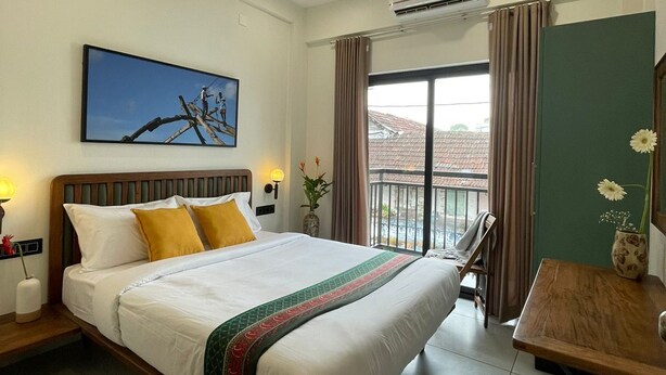 A hotel room with a bed, a desk, a chair, an air conditioner, a window with curtains, and a painting on the wall.