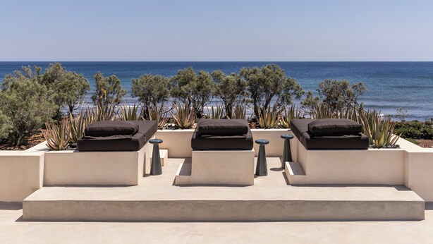 Un salón junto al mar con tres zonas de estar acolchadas, cada una con un cojín, rodeado de plantas y vistas al océano.