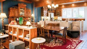 A cozy room with a wooden dining table, chairs, a chandelier, and a kitchen area with cabinets and a window.