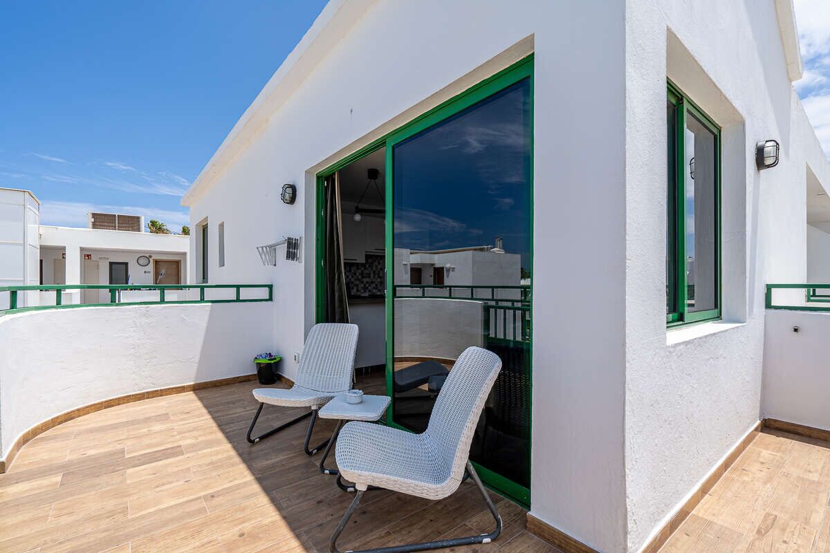 Un balcon avec deux fauteuils en osier et une petite table, une porte vitrée, et un bâtiment blanc doté d’une bordure verte.