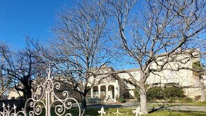 A courtyard with a white wrought iron fence, a building with arches, and a tree in the background. - demi-château de Paulignan for 15 people (TRAUSSE)