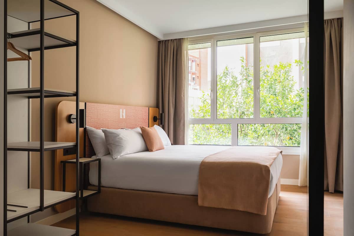 A modern hotel room with a large bed, a built-in wardrobe, and a window with a view of greenery.