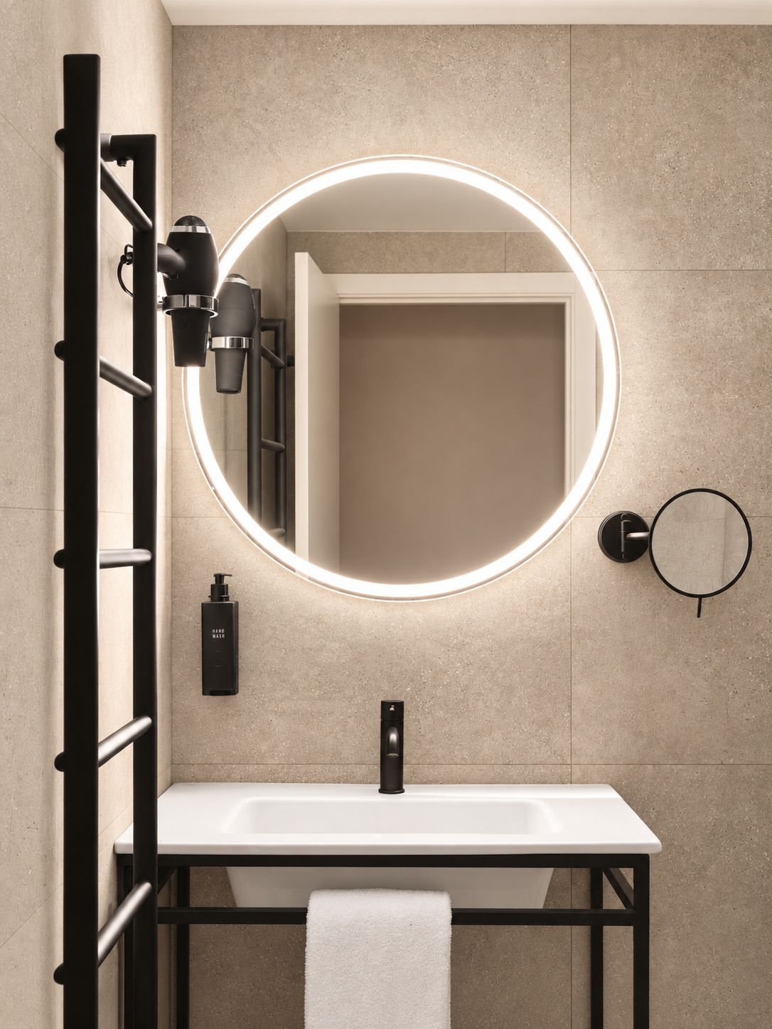 Un baño moderno con un lavamanos blanco, un espejo redondo con luz y una toalla colgada en un perchero.