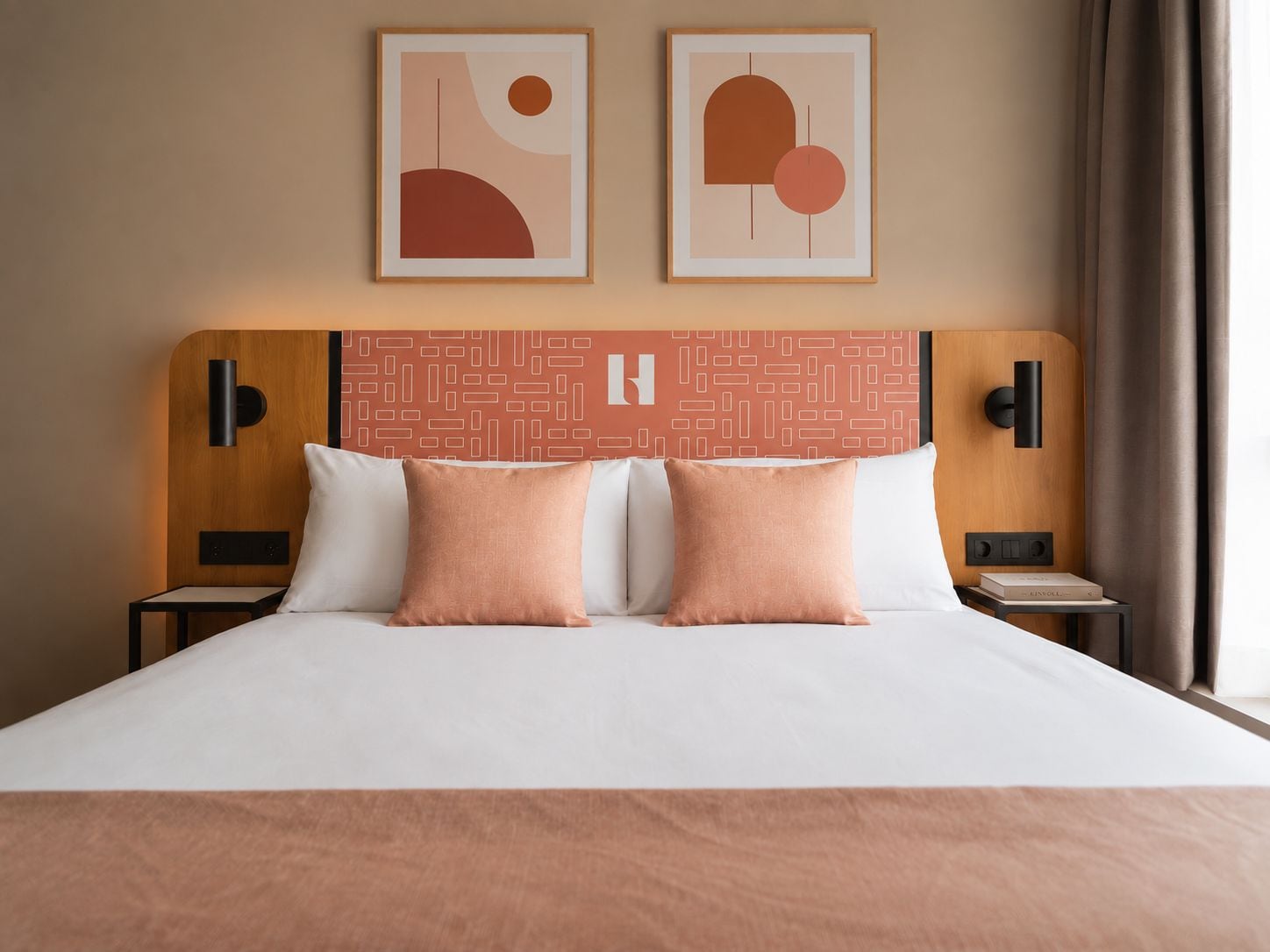 A hotel room with a large bed, two pillows, a headboard with a geometric pattern, two bedside tables with lamps, and two framed abstract artworks on the wall.