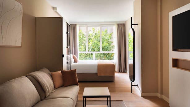A modern hotel room with a sofa, a bed, a TV, and a window with curtains. - Limehome Bilbao Deusto (Bilbao)