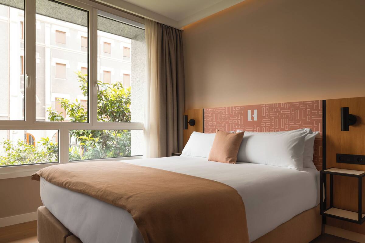 A hotel room with a large bed, a view of buildings and greenery outside, a wooden headboard, and a small bedside table.