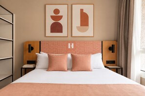 Ein Hotelzimmer mit einem Bett, zwei Kissen, einem Kopfteil mit geometrischem Muster, einem Wandbild und einem Nachttisch mit Lampe.