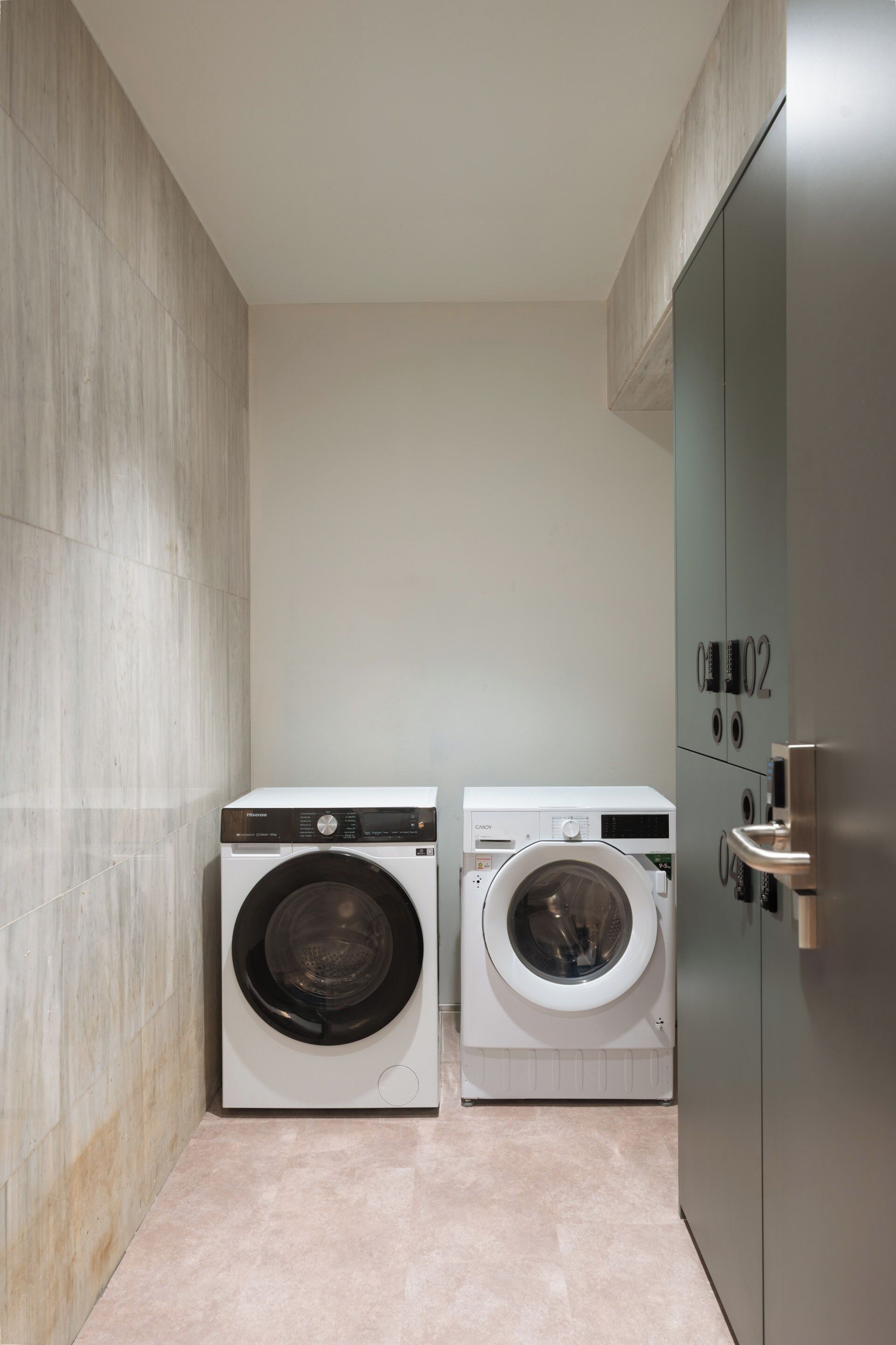A compact laundry room with two front-loading washing machines, a tiled floor, and a metallic door with a number plate.