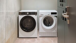 A compact laundry room with two front-loading washing machines, a tiled floor, and a metallic door with a number plate. - Limehome Bilbao Deusto (Bilbao)