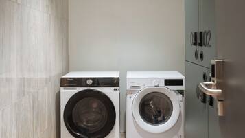 A compact laundry room with two front-loading washing machines, a tiled floor, and a metallic door with a number plate.