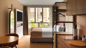 A hotel room with a bed, a desk with a laptop, a chair, a TV, and a window with a view of buildings and greenery. - Limehome Bilbao Deusto (Bilbao)
