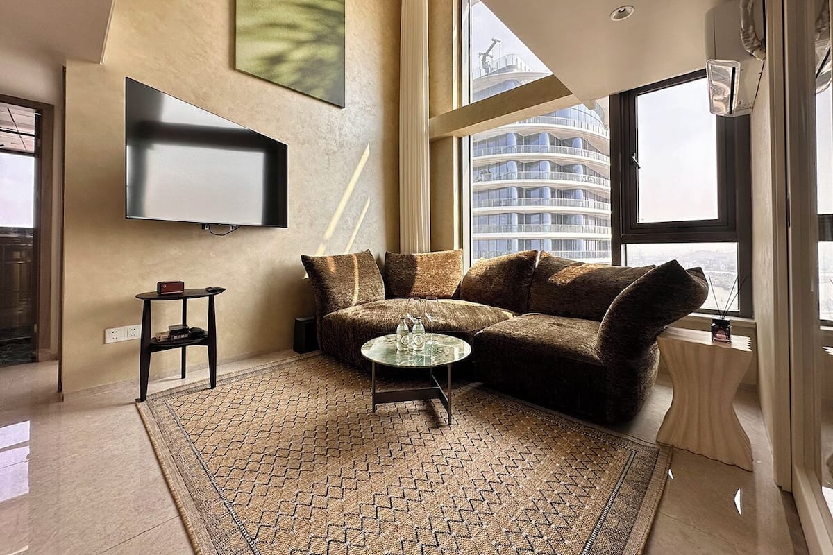 Una sala de estar moderna con un sofá seccional, una mesa de centro de vidrio y un ventanal que ofrece vistas a un edificio de rascacielos.
