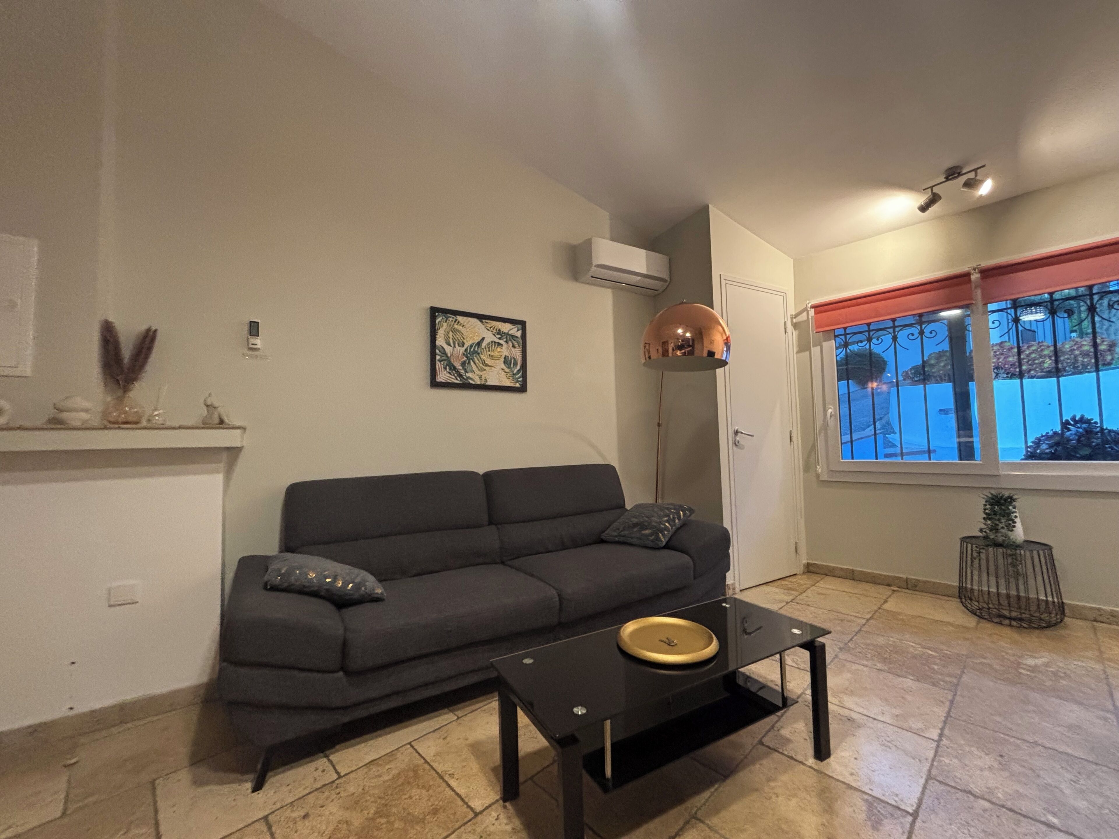 A living room with a sofa, a coffee table, a picture frame, an air conditioning unit, and a window with a view.