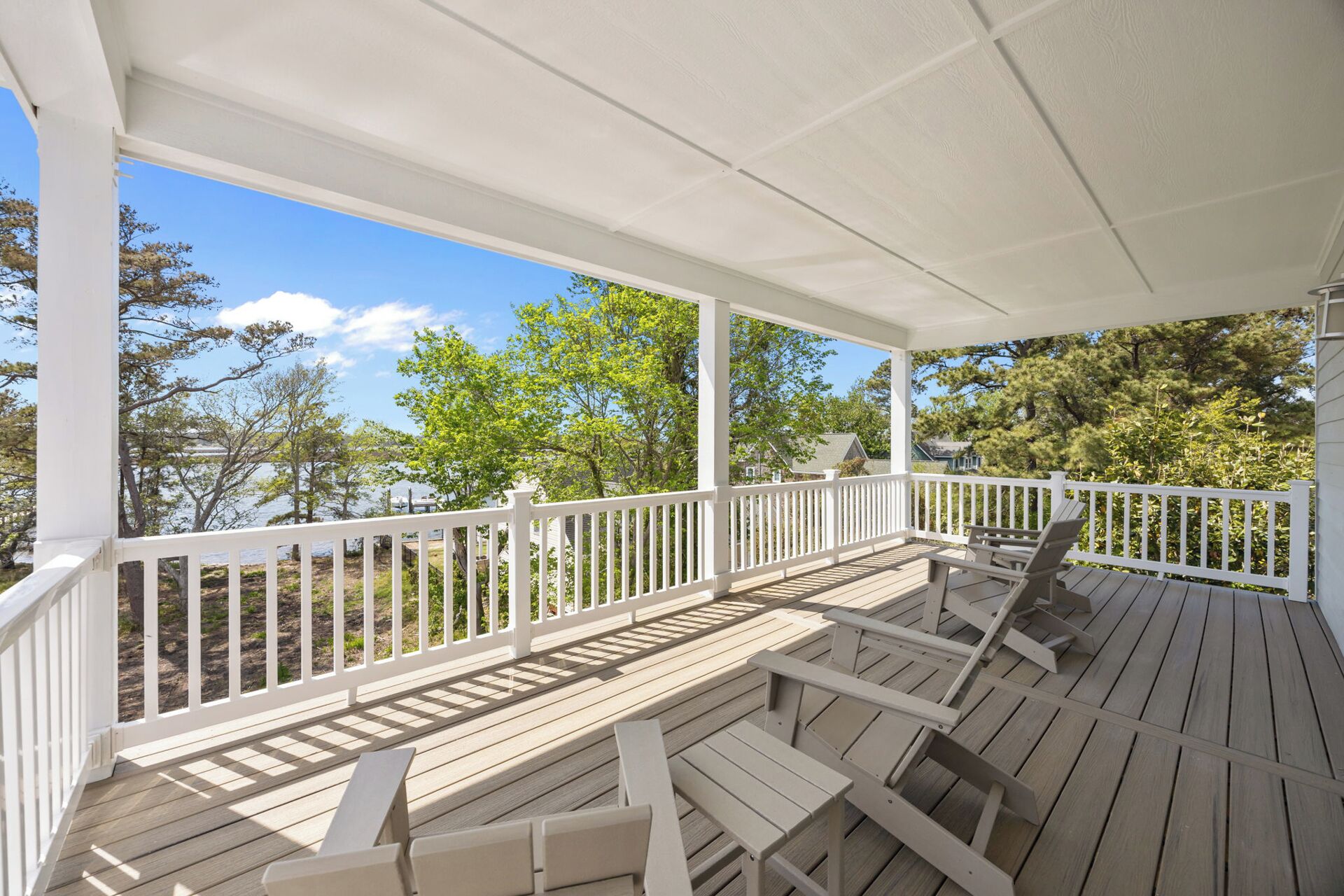 A covered wooden deck with white railings, outdoor seating, and a view of trees and a body of water.