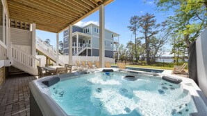 A hot tub under a wooden pergola, a poolside area with lounge chairs, and a multi-story building in the background. - BRAND NEW Saltaire 8 Bedroom Luxury Home at Water Oak (Kill Devil Hills)