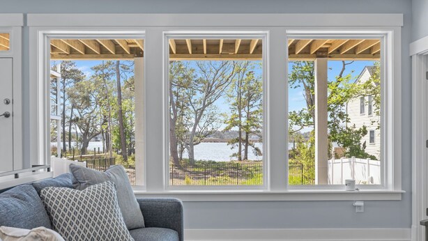 A room with a large window, a sofa, a coffee table, and a view of a lake and trees. - BRAND NEW Saltaire 8 Bedroom Luxury Home at Water Oak (Kill Devil Hills)