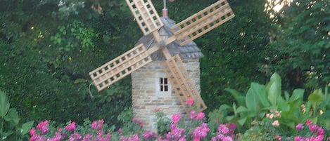 Un molino de piedra con estructura de celosía, rodeado por un muro de piedra y vibrantes flores rosadas.