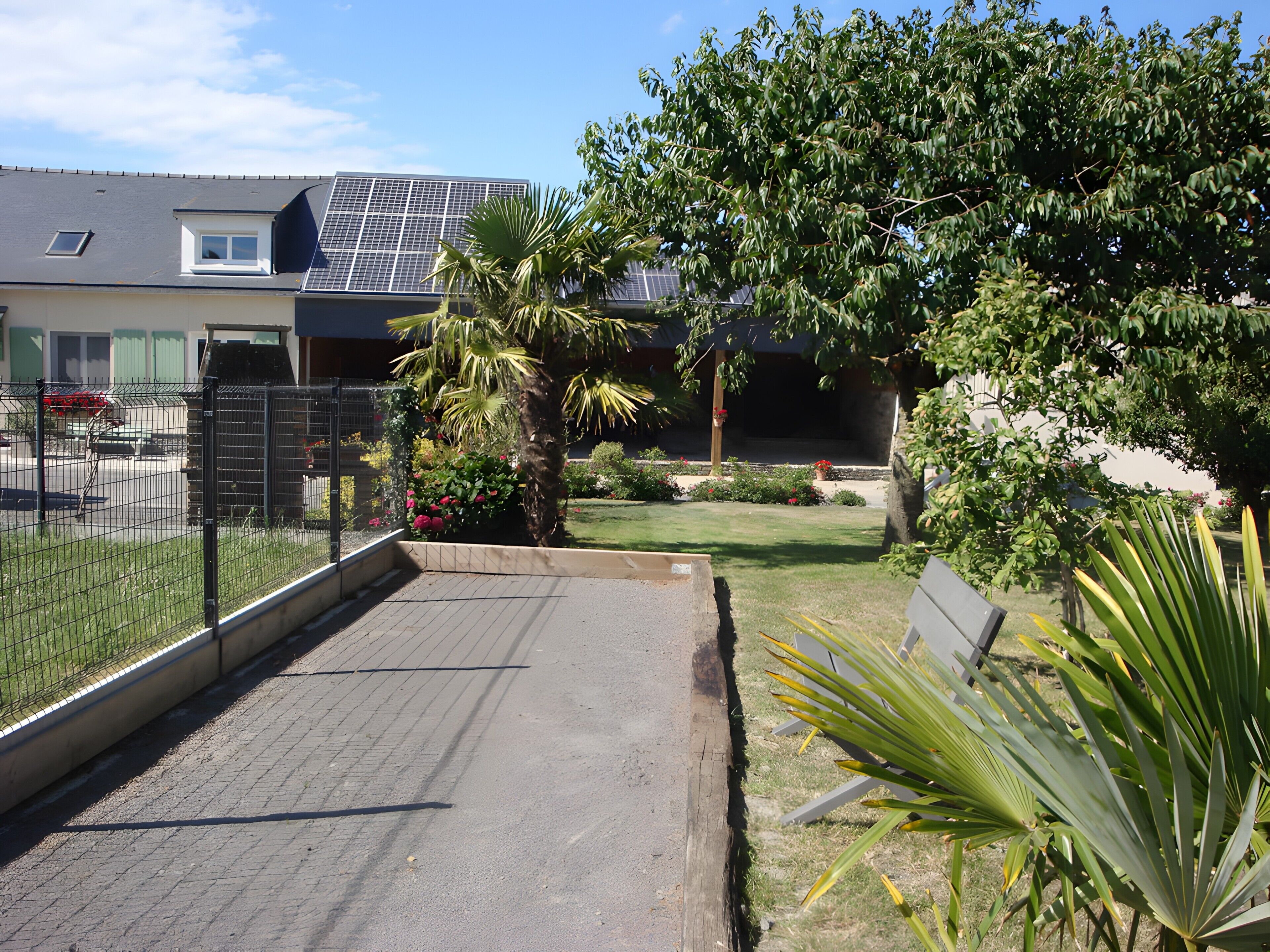 Un chemin pavé mène à un bâtiment dont le toit est recouvert de panneaux solaires, et qui est entouré de verdure et d’une zone délimitée par une clôture.