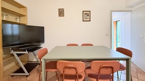 Ruokailutila, jossa on pöytä ja tuolit, jalustalla oleva televisio sekä kaksi kehystettyä kuvaa seinällä.