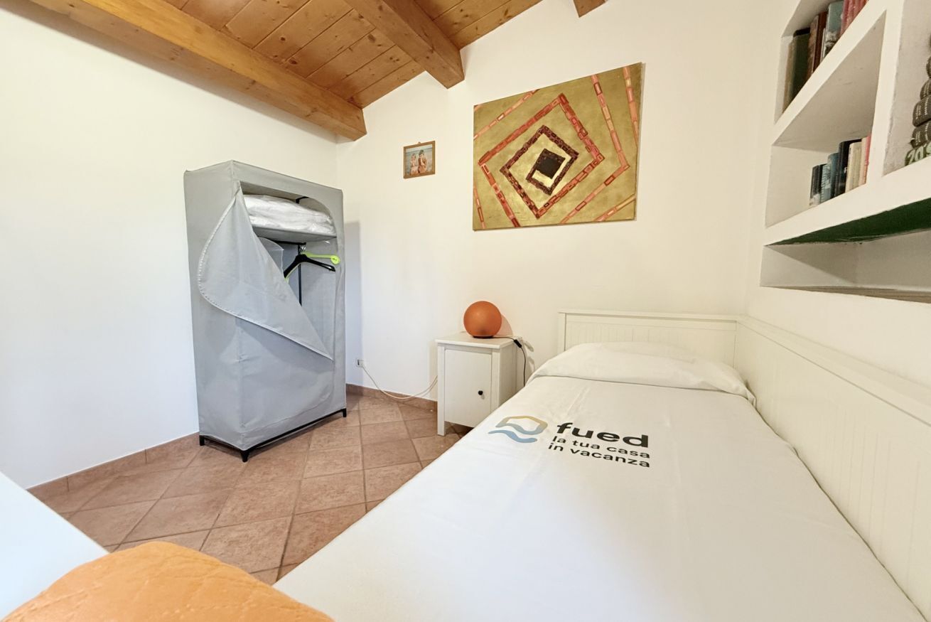 A hotel room with a bed, a wardrobe, a small table with a lamp, a painting on the wall, and a bookshelf.