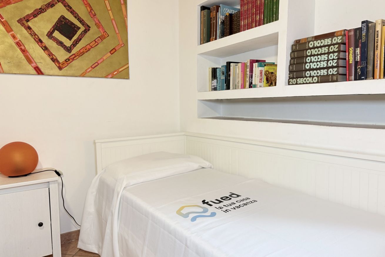 Una cama individual con una colcha blanca, una estantería llena de libros, una mesita de noche con una lámpara y un adorno en la pared.