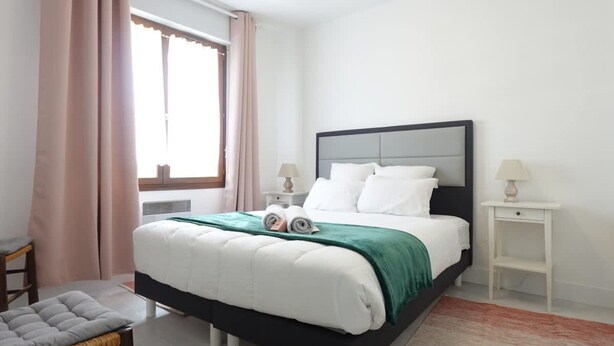 Un dormitorio con cama, una silla, una mesita de noche y una ventana con cortinas.