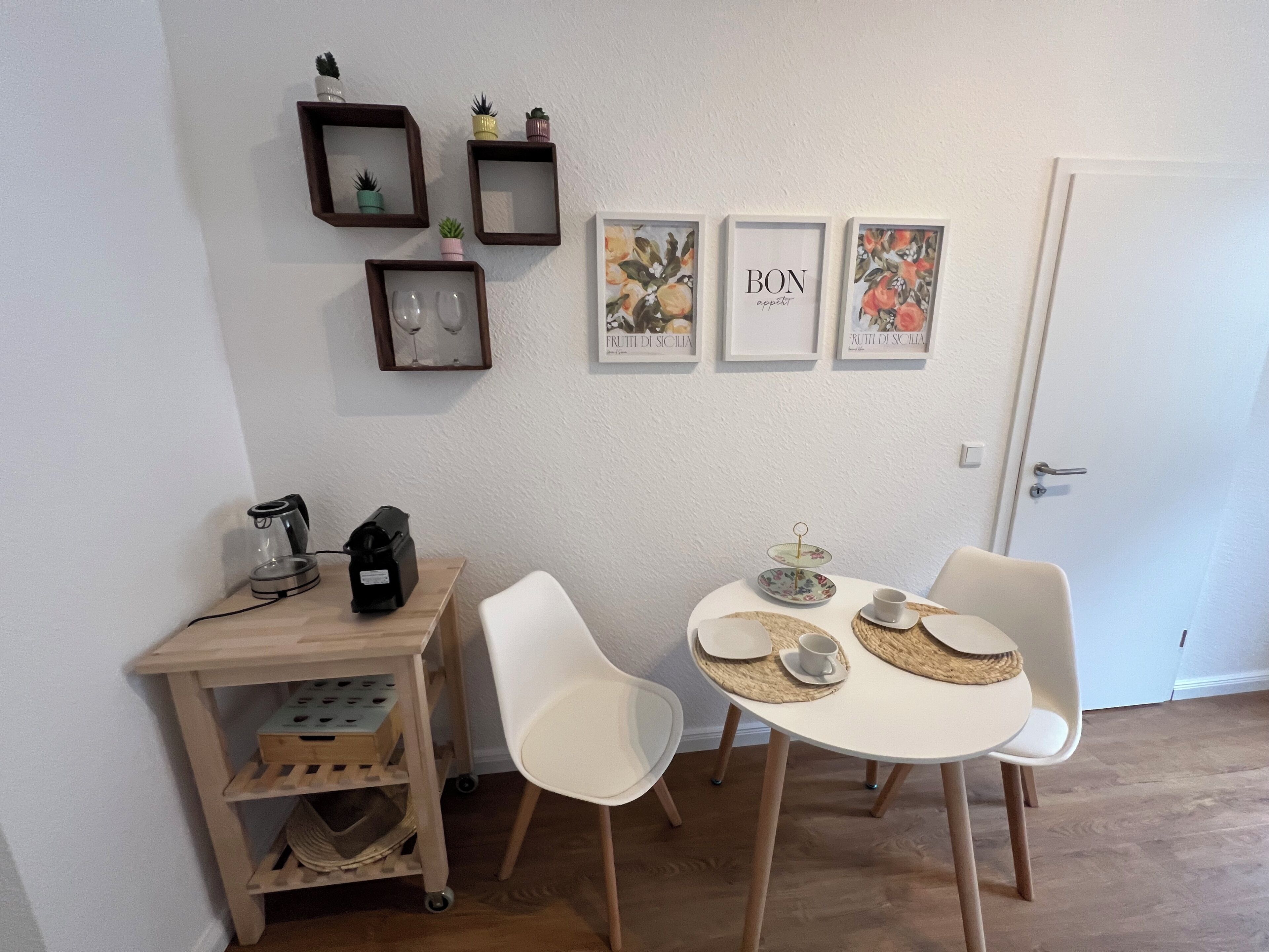 En matplats med ett vitt bord, två stolar, en kaffemaskin, en liten hylla med dekorativa föremål och inramade tavlor på väggen.