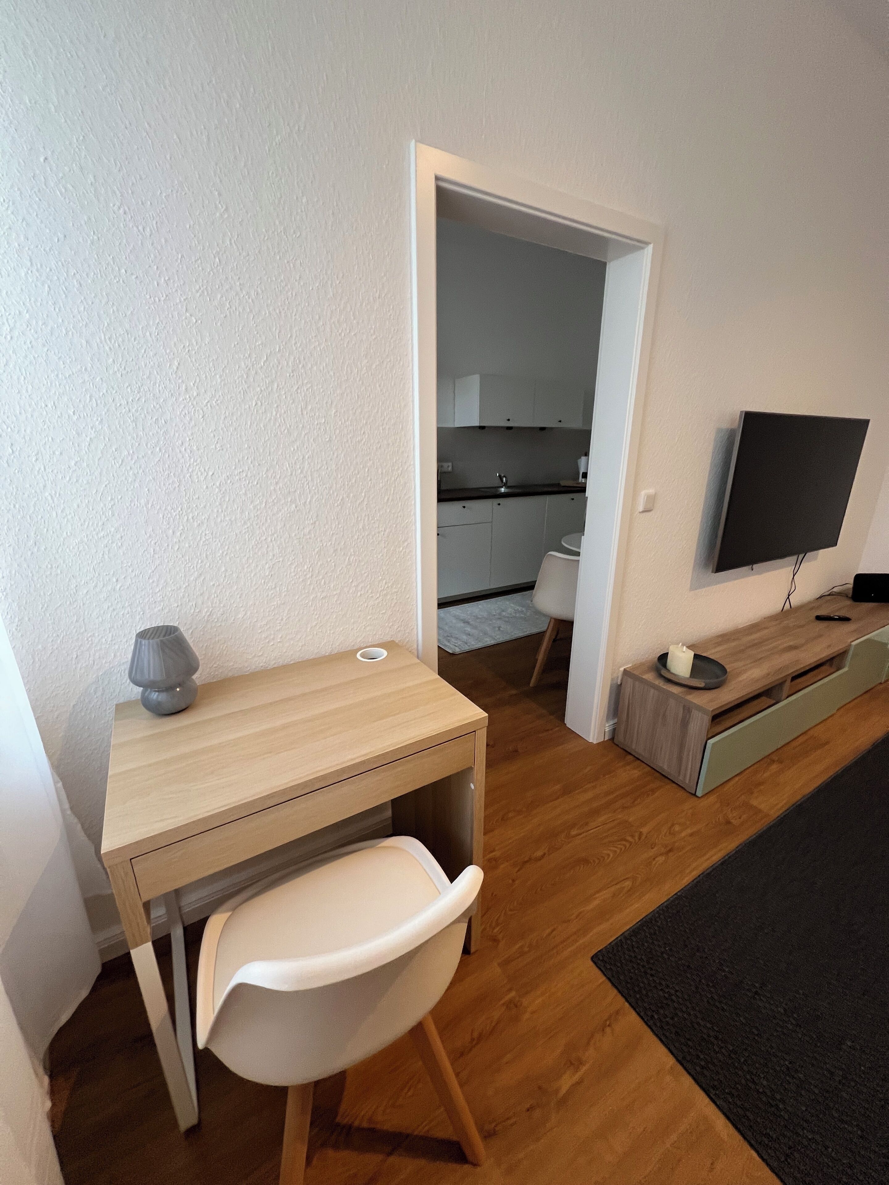 Une petite pièce moderne, dotée d’un bureau en bois, d’une chaise blanche, d’un téléviseur fixé au mur et d’une cuisine visible à travers une porte ouverte.