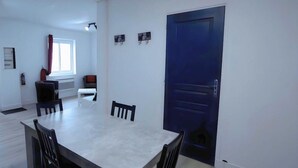 Ein Zimmer mit Esstisch, Stühlen, einem Fenster mit Vorhängen, einer blauen Tür und einem kleinen Kleiderständer mit Gegenständen.