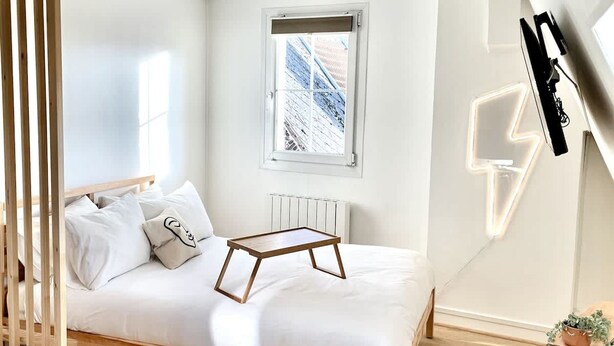 A minimalist bedroom with a wooden bed, a window with a view, a radiator, and a wall-mounted light fixture. - Appartement Lumineux Scandinave - Coup de Foudre (Arras)
