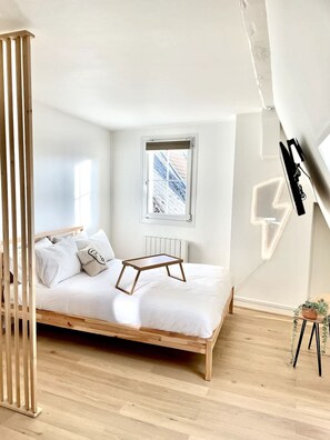 A minimalist bedroom with a wooden bed, a window with a view, a radiator, and a wall-mounted light fixture. - Appartement Lumineux Scandinave - Coup de Foudre (Arras)