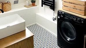 A modern bathroom with a built-in washing machine, a black towel rack, a window, and a patterned floor. - Appartement Lumineux Scandinave - Coup de Foudre (Arras)