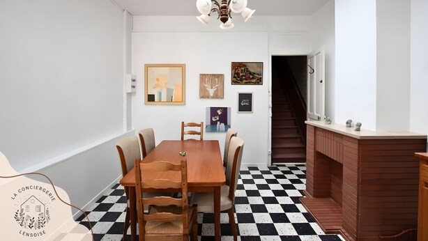 Ein Essbereich mit Schachbrettboden, einem Holztisch und Stühlen. An den Wänden befinden sich gerahmte Kunstwerke und eine Treppe, die zu einer anderen Ebene führt.