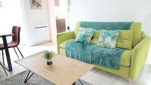 Ein grünes Sofa mit einem türkisfarbenen Überwurf und tropischen Kissen, ein Couchtisch aus Holz mit einer Pflanze und ein gemustertes Teppich.