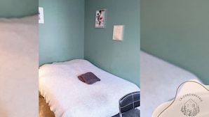 Uma cama de solteiro com cobertura branca e travesseiro marrom, uma pequena mesa de cabeceira e um interruptor de luz fixado na parede.