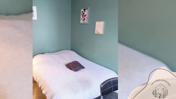 Uma cama de solteiro com cobertura branca e travesseiro marrom, uma pequena mesa de cabeceira e um interruptor de luz fixado na parede.